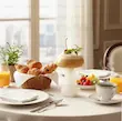 لیوان شیشه‌ای طرح قارچ با نوشیدنی پرتقالی و فوم سفید روی میز صبحانه کنار کروسان و فنجان قهوه