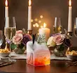 لیوان شیشه‌ای دوجداره شفاف طرح پاکت شیر با نوشیدنی نارنجی و کف سفید، وسط میز شام نورانی