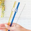 دست در حال نگه داشتن دو نی شیشه‌ای خمره‌ای طلایی و آبی همراه برس تمیزکننده