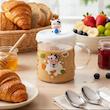 ماگ شیشه‌ای فانتزی طرح گاو با نوشیدنی گرم روی میز صبحانه، کنار کروسان و تزیینات رنگی