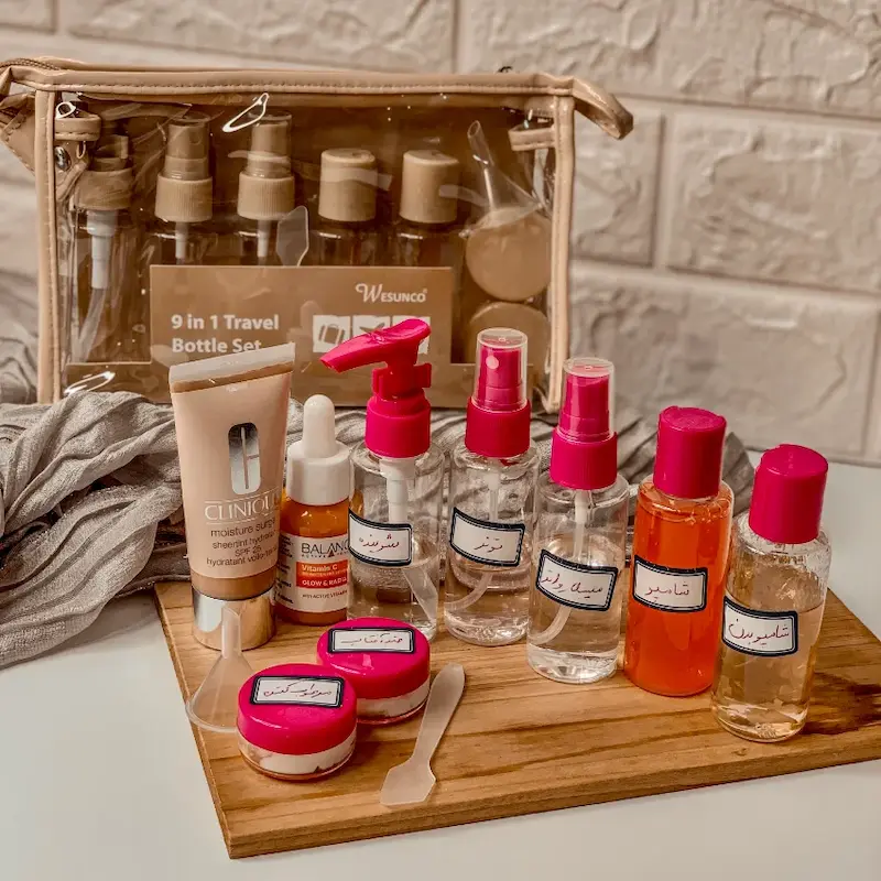 پک مسافرتی بهداشتی ۹ تیکه با بطری‌های اسپری و کرم‌دان صورتی داخل کیف کرمی شفاف روی میز چوبی