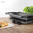 گریل بوش مدل TCG4215 در حال پرس کردن ساندویچ پانینی روی میز چوبی آشپزخانه در کنار گلدان.