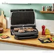 گریل باربیکیو تفال مدل GC722D روی میز چوبی آشپزخانه در حال گریل کردن استیک و سوسیس. 