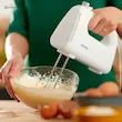 یک فرد در حال مخلوط کردن خمیر کیک در کاسه شیشه ای با همزن دستی فیلیپس مدل HR3705 سفید.
